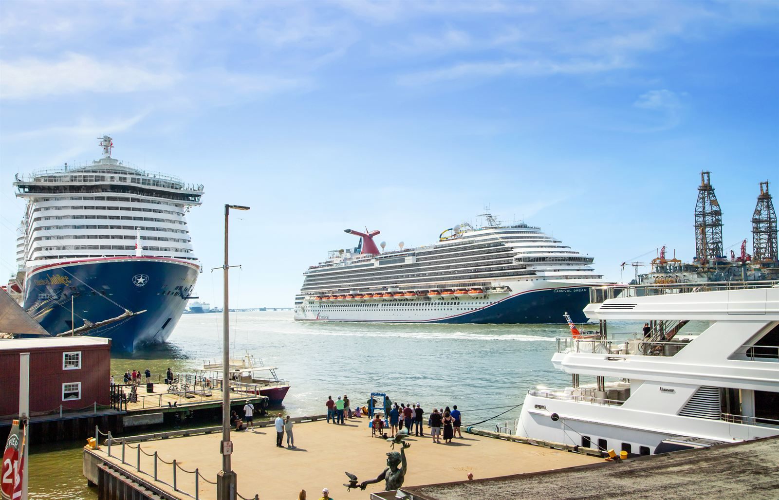 Two cruise ships in the Galveston harbor channel seen from a waterfront pier with people and a yacht