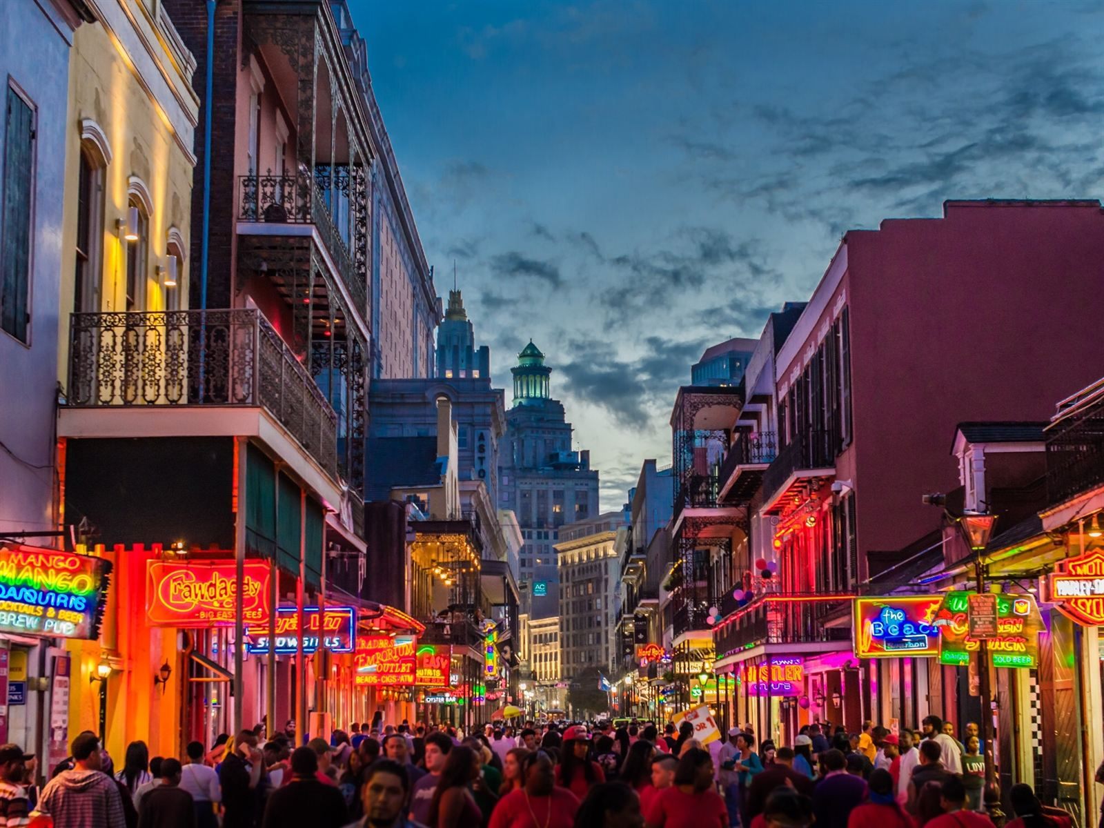 Crowds on Bourbon Street in the French Quarter at dusk under neon signs and iron balconies