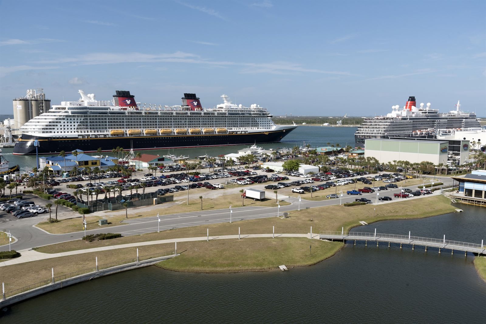 Two cruise ships docked at Port Canaveral behind a parking area, lagoon and footbridge